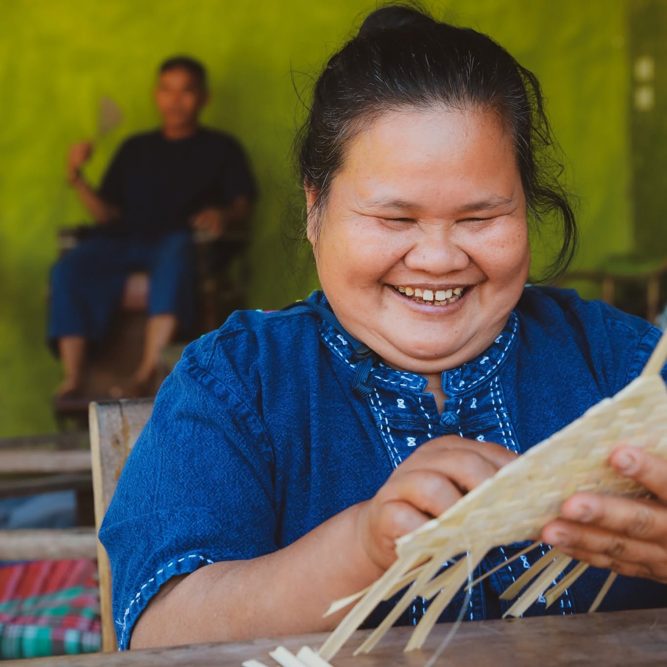 Aunt Thong smiles while weaving a bamboo fan with a guest